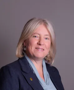 Eine Frau mit blonden Haaren lächelt und schaut in die Kamera. Sie trägt einen marineblauen Blazer, ein graues Hemd und eine Anstecknadel an ihrem Blazer.