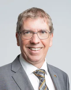 Ein lächelnder Mann mit Brille, der einen grauen Anzug und eine karierte Krawatte trägt. Er sieht geradeaus und befindet sich wahrscheinlich in einer professionellen Umgebung.
