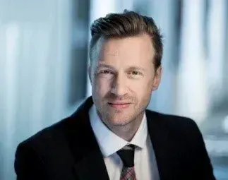Ein Mann in einem schwarzen Anzug und Krawatte lächelt in die Kamera. Er hat kurze Haare und steht in einem gut beleuchteten Raum.