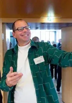 Ein Mann in einem grünen Jackett mit Namensschild lächelt und zeigt Daumen hoch in einem Flur mit anderen Leuten.