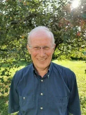 Ein älterer Mann mit Brille und blauem Hemd steht auf einem Feld, mit einem großen Baum hinter ihm. Er hat ein breites Lächeln im Gesicht.