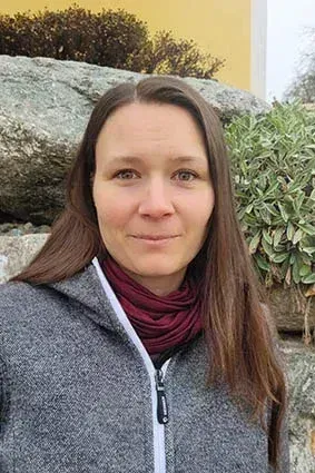 Eine Frau mit langen braunen Haaren und einem roten Schal lächelt vor einer Felswand mit Grünpflanzen. Sie trägt eine graue Kapuzenjacke mit Reißverschluss.