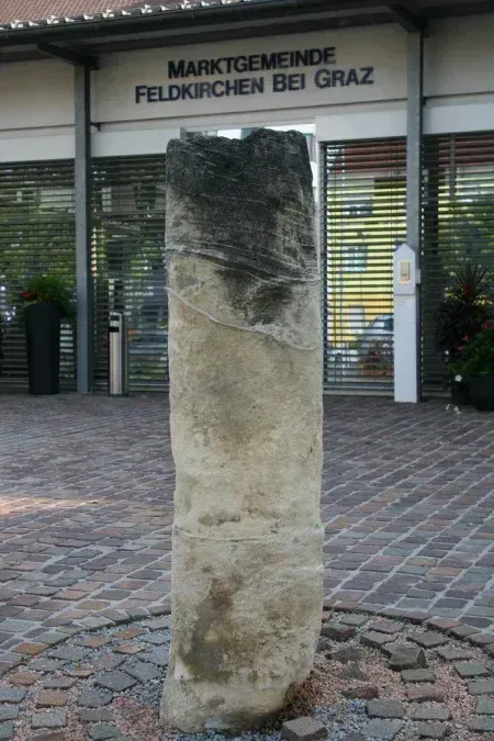 Ein alter Steinpfeiler steht vor einem Gebäude mit einem Schild, das Marktgemeinde Feldkirchen bei Graz liest. Der Pfeiler ist von einem Kopfsteinpflaster umgeben und es gibt Topfpflanzen in der Nähe.
