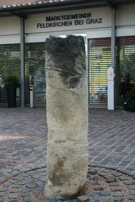 Ein alter Steinpfeiler steht vor einem Gebäude mit einem Schild, das Marktgemeinde Feldkirchen bei Graz liest. Der Pfeiler ist von einem Kopfsteinpflaster umgeben und es gibt Topfpflanzen in der Nähe.