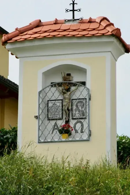 Eine kleine Kapelle mit einem Kreuz, einem Kreuz oben und einem Blumentopf unten. An der Wand hängen zwei Bilder.