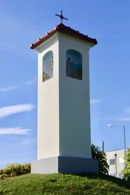 Ein weißer Turm mit einem Kreuz auf der Spitze steht auf einem Grashügel, mit zwei Gemälden religiöser Figuren an der Seite.