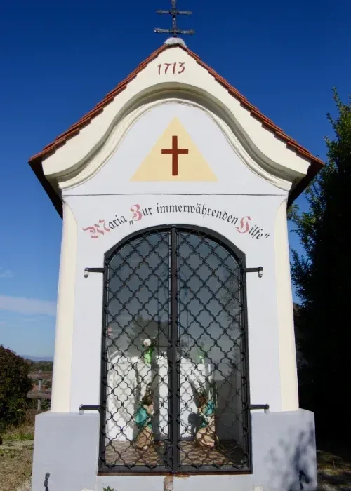 Eine kleine Kapelle mit einem Kreuz auf der Spitze, datiert 1713. Die Kapelle hat eine Glastür mit Metalldesign, und im Inneren befinden sich zwei Statuen mit Blumen. Die Wand hat eine deutsche Inschrift.