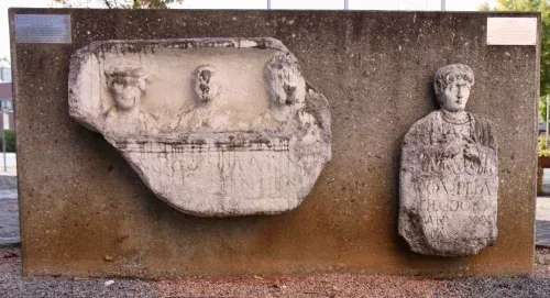 Eine Steintafel mit einer geschnitzten Darstellung von vier Figuren, montiert an einer Betonwand. Daneben befindet sich eine kleinere geschnitzte Figur.