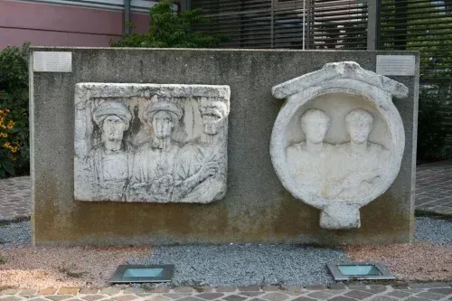 Eine Betonwand mit zwei gemeißelten Steinplatten, eine zeigt vier Personen und die andere zwei Gesichter, umgeben von Pflanzen und Kies.