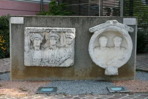 Eine Betonwand mit zwei gemeißelten Steinplatten, eine zeigt vier Personen und die andere zwei Gesichter, umgeben von Pflanzen und Kies.