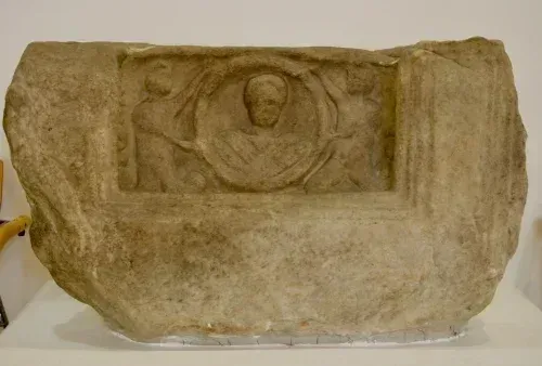 Eine antike Steinskulptur, die ausgestellt ist, mit einem geschnitzten Relief einer zentralen Figur, umgeben von zwei kleineren Figuren. Die Skulptur hat ein abgenutztes Aussehen, mit sichtbaren Rissen und Absplitterungen.