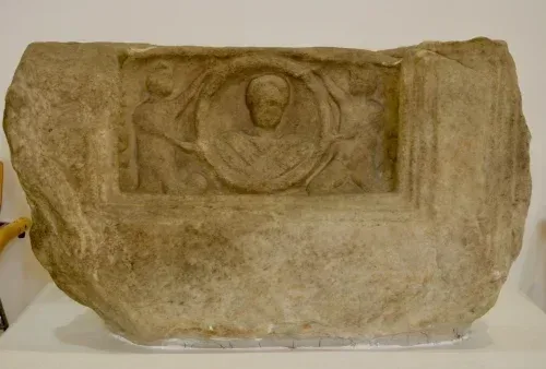 Eine antike Steinskulptur, die ausgestellt ist, mit einem geschnitzten Relief einer zentralen Figur, umgeben von zwei kleineren Figuren. Die Skulptur hat ein abgenutztes Aussehen, mit sichtbaren Rissen und Absplitterungen.