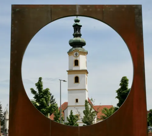 Ein hoher Glockenturm ist durch eine kreisförmige Öffnung in einem braunen Metallrahmen zu sehen. Der Turm hat ein grünes Dach und eine Uhr. Bäume und ein Gebäude mit einem roten Dach sind im Hintergrund sichtbar.