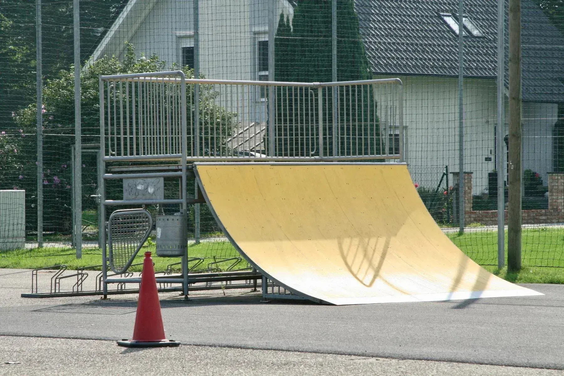 Eine gelbe Rampe, möglicherweise zum Skateboarden, steht neben einem Metallgeländer. Ein roter Verkehrskegel steht in der Nähe. Hinter der Rampe befindet sich ein Gebäude mit einem Maschendrahtzaun und Bäumen.