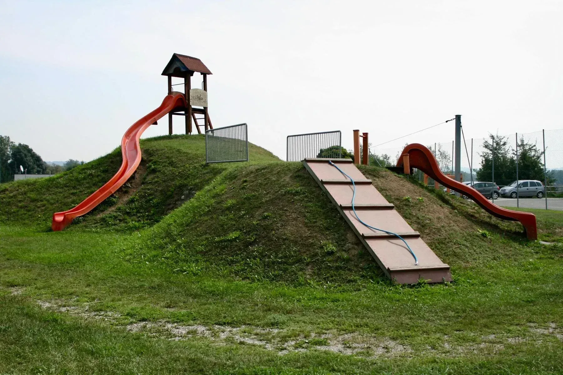 Ein Freiluftspielplatz mit einer roten Rutsche, einem Aussichtsturm und einem eingezäunten Bereich auf einem Grashügel.