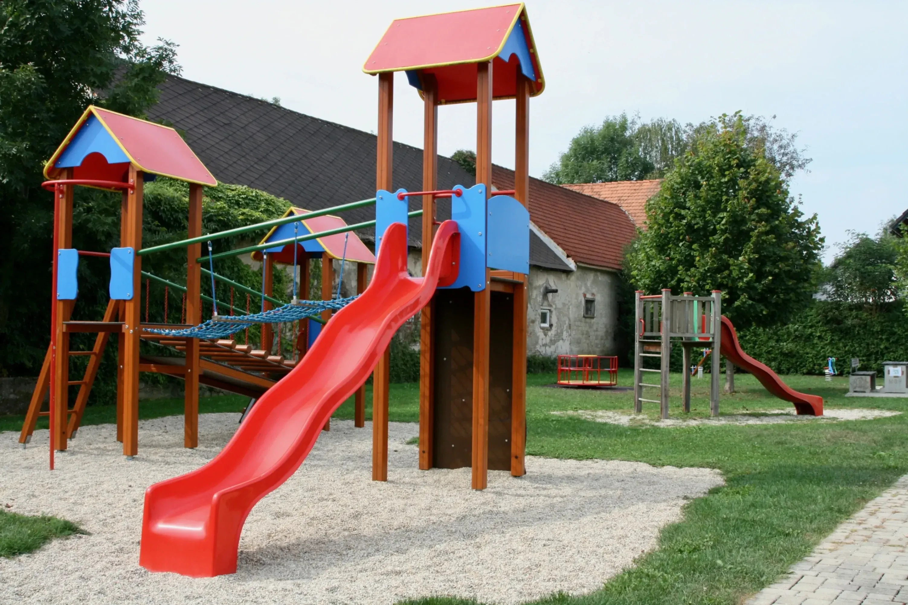 Ein bunter Spielplatz mit einer roten Rutsche, einem Klettergerüst und einem kleinen Haus mit rotem Dach im Hintergrund.