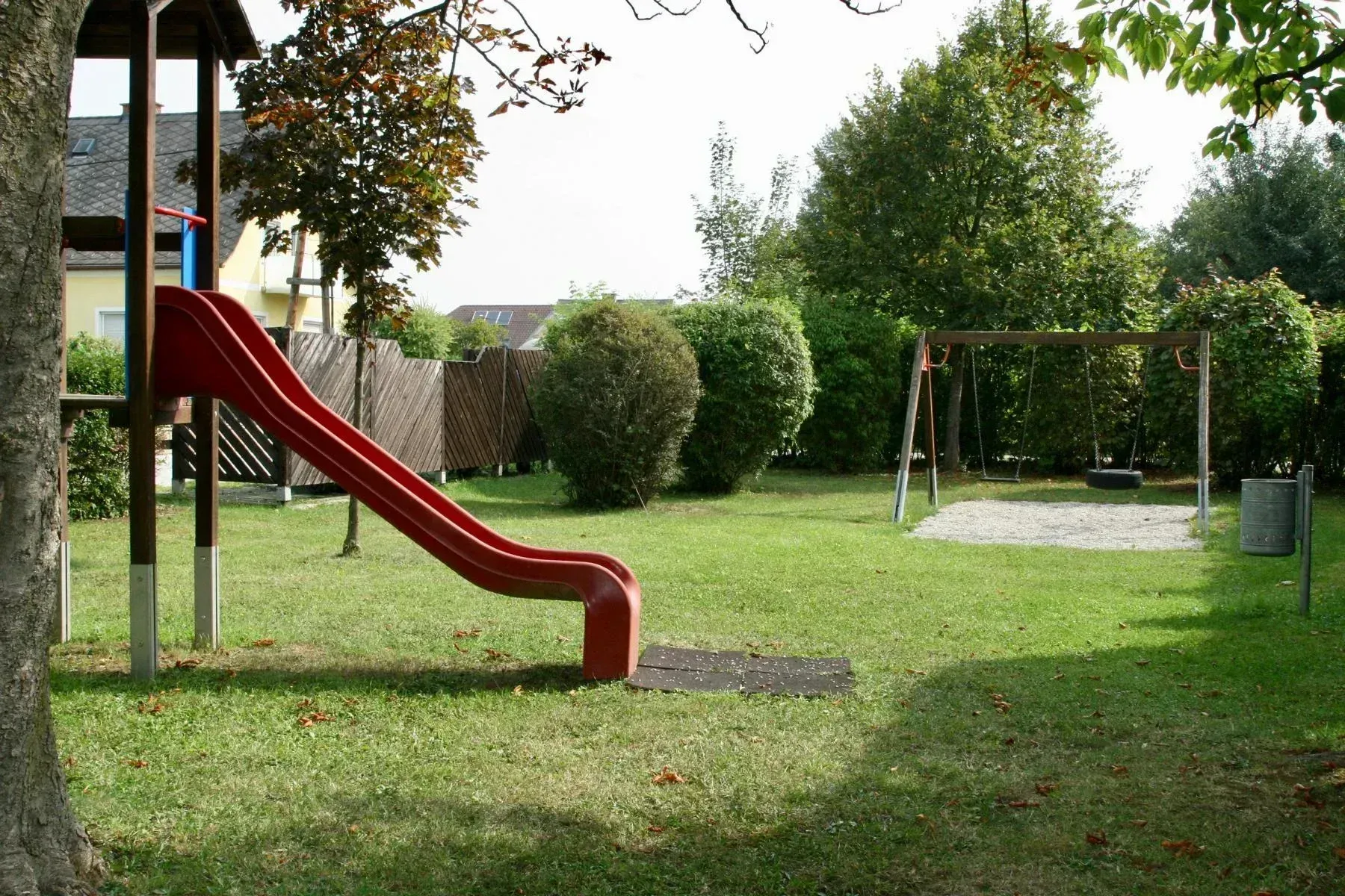 Ein Garten mit einem roten Rutschen und einer Schaukel, umgeben von Grün. Ein Zaun und ein Haus sind im Hintergrund.