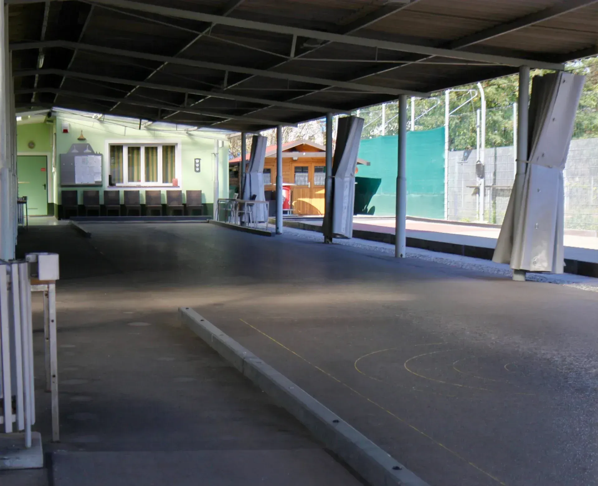 Ein leerer Sportplatz mit Dach, einer Wand und einigen Bänken. Es gibt ein Gebäude mit einem Fenster und einem Mülleimer in der Nähe.