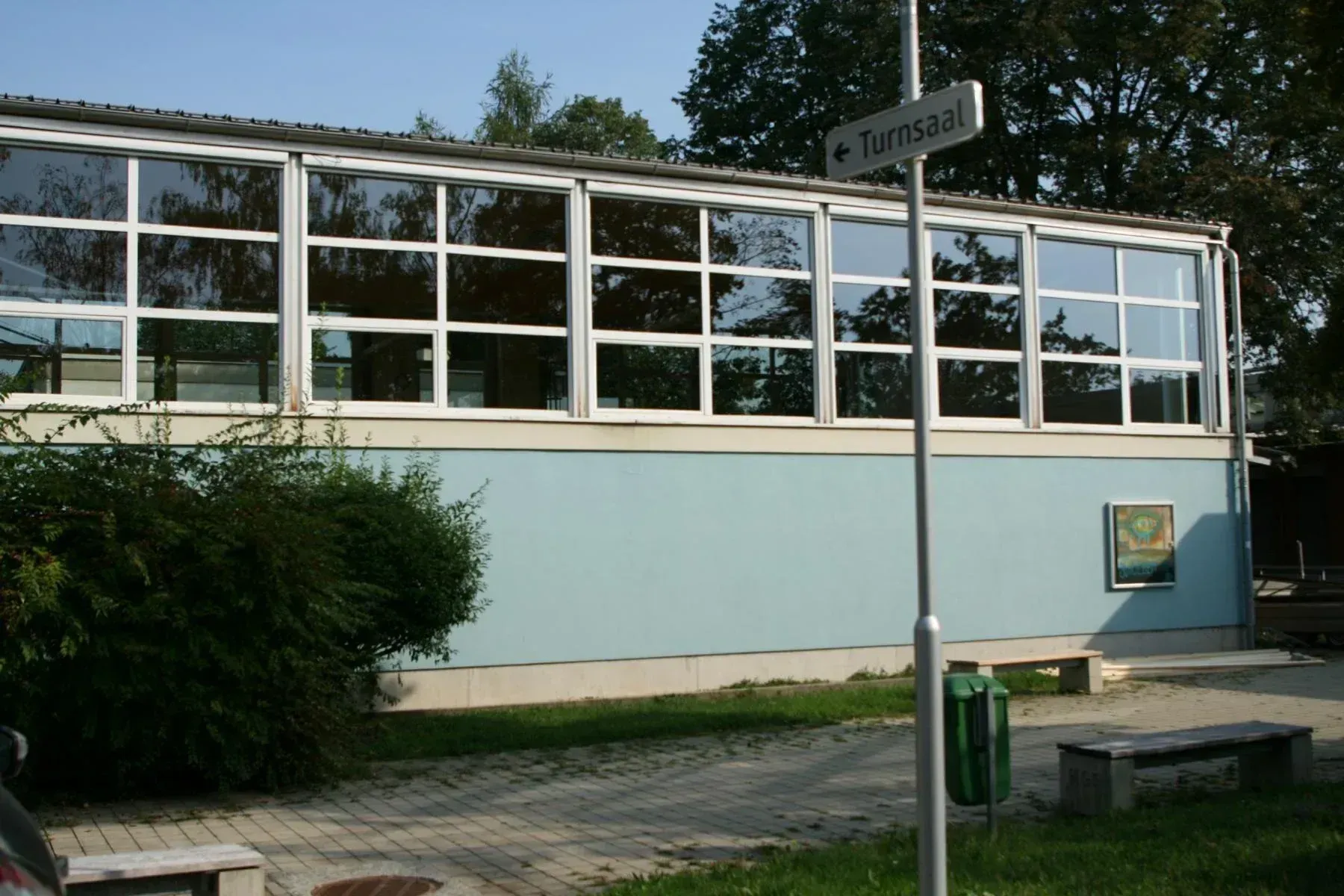 Ein Gebäude mit vielen Glasfenstern und einem Schild für Turnsaal. Bäume und eine Bank sind im Hintergrund.