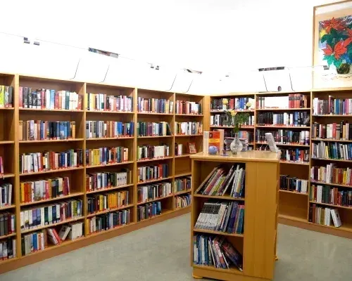 Eine gut organisierte Bibliothek mit zahlreichen Holzregalen voller Bücher. Ein kleiner Tisch mit Büchern und einer Blumenvase steht in der Mitte. Der Raum ist gut beleuchtet.