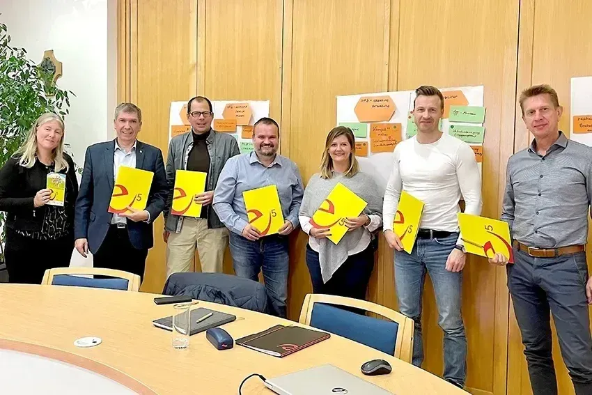 Eine Gruppe von Menschen steht in einem Konferenzraum, hält gelbe Papiere mit Zahlen und lächelt für ein Foto. Hinter ihnen hat eine Wand ein Whiteboard und farbige Haftnotizen. Ein Tisch hat einen Laptop, eine Maus und ein Notizbuch.