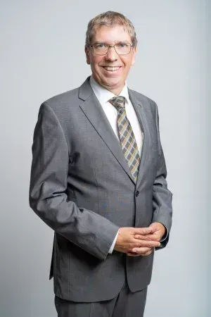 Ein Mann in einem grauen Anzug und mit Brille lächelt und hat die Hände vor sich verschränkt.