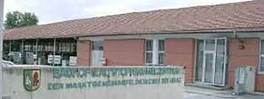 Ein Gebäude mit einem roten Dach und Ziegelwänden. Ein Schild vor dem Gebäude lautet 'SPARCHE KANTOY PARMALISTION'. Das Gebäude hat mehrere Fenster und ein kleines Logo auf der linken Seite.
