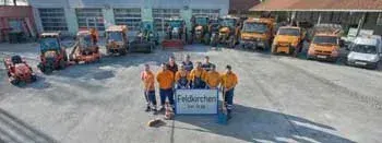 Eine Gruppe von Menschen in orangefarbenen Uniformen posiert für ein Foto vor einem Schild, das 'Feldkirchen' steht. Im Hintergrund sind zahlreiche Baufahrzeuge und Lkws auf einem Parkplatz geparkt.