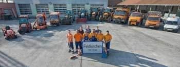 Eine Gruppe von Menschen steht vor einem Schild mit Text, umgeben von verschiedenen Baumaschinen, darunter Bagger, Lastwagen und ein Gabelstapler, in einem Außenbereich.