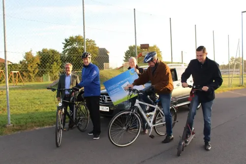 Fünf Männer sind draußen mit Fahrrädern und Rollern. Ein Mann hält ein großes blaues Schild. Der Mann in der Mitte hält ein Schild mit einem Auto hinter ihm.