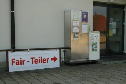 Ein Metallbehälter mit einer Glastür und mehreren Plakaten steht draußen. Ein Schild mit dem Wort Weiler steht auf dem Bürgersteig.