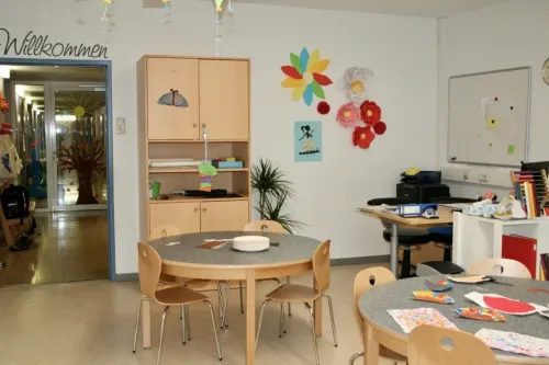 Ein Klassenzimmer mit einem runden Tisch und Stühlen, einem Holzschrank, einer Tafel und bunten Blumendekorationen an der Wand.