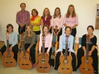 Eine Gruppe von Personen, möglicherweise eine Band, posiert für ein Foto mit ihren Gitarren. Sie sitzen auf Stühlen, einige stehen, alle lächeln und schauen nach vorne. Der Raum hat eine weiße Wand und ein Bücherregal.