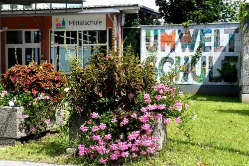 Das Äußere eines Schulgebäudes mit dem Schild 'Mittelschule'. An der Wand befindet sich ein Wandbild mit dem Wort 'Umweltschule'. Vor dem Gebäude steht eine Pflanze mit rosa Blüten.