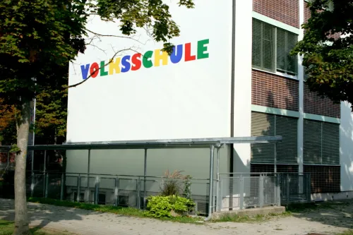 Ein weißes Gebäude mit dem Wort Volksschule in mehrfarbigen Buchstaben bemalt. Das Gebäude hat Metallgeländer und einen Baum davor.