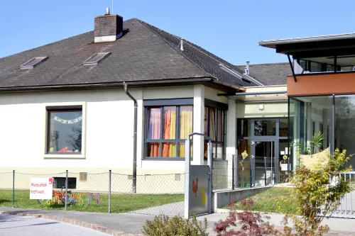 Ein einstöckiges Gebäude mit grauem Dach und Schornstein. Es hat Glasfenster mit Vorhängen und eine Glastür. Der Eingang hat ein Tor mit einem Hahnenbild. Vor dem Gebäude steht ein Zaun.