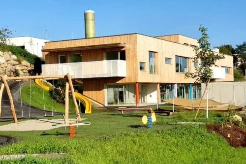 Ein modernes Gebäude mit Balkon und Schornstein, umgeben von einem Spielplatz mit Rutsche und Schaukel, und einem Rasenbereich mit Bank und Baum.
