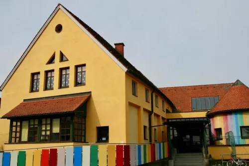 Ein leuchtend gelbes Gebäude mit mehreren Fenstern und einem roten Dach. Der Eingang befindet sich auf der rechten Seite mit einer Treppe, die hinaufführt. Vor dem Gebäude steht ein farbenfroher Zaun.