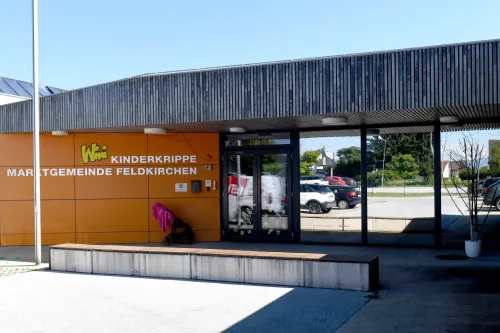 Ein orangefarbenes Gebäude mit den Worten Kinderkrippe Inde Feldkirchen, ein Kinderwagen davor und Autos in der Ferne geparkt.