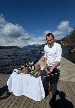 Ein Mann in Kochkleidung bereitet ein Essen am Seeufer zu, umgeben von Flaschen und einem Tisch mit Essen.
