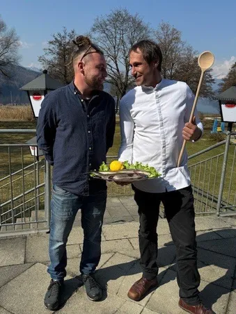 Zwei Männer stehen in einem Park mit Bergen im Hintergrund. Ein Mann hält einen Holzlöffel und einen Teller mit Essen, der andere Mann hat ein lächelndes Gesicht.