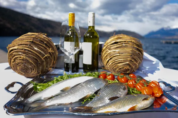 Ein Tisch mit Fisch, Weinflaschen und Tomaten am Ufer eines Bergsees. Zwei Weidenkörbe stehen auf beiden Seiten des Tisches.