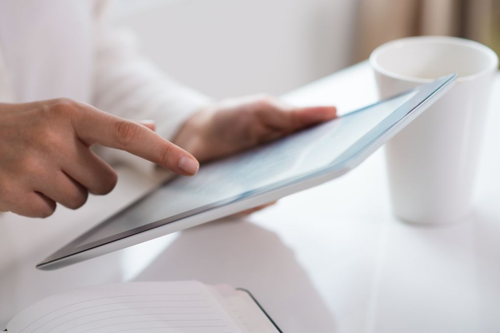  Abschnitt Bild: Eine Person zeigt mit dem Finger auf einen Tablet-Bildschirm, daneben liegt ein offenes Notizbuch. Ein Becher steht auf einem Tisch im Hintergrund.