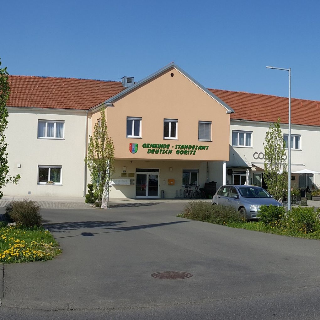  Abschnitt Bild: Die Vorderansicht eines Gebäudes mit einem roten Dach und den Worten Gemeinde-Standesamt Deutsch Goritz. Vor dem Gebäude ist ein Auto geparkt.