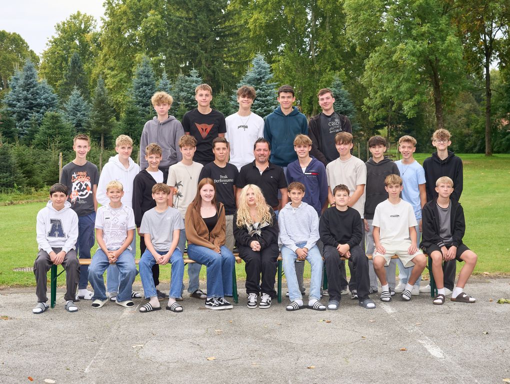  Abschnitt Bild: Eine Gruppe junger Menschen, möglicherweise Schüler, versammelt sich für ein Klassenfoto in einem Park. Sie sitzen auf Bänken, und einige stehen. Bäume sind im Hintergrund.