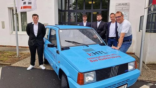 Fünf Männer stehen vor einem blauen Auto, zwei tragen eine Brille und einer hat eine Uhr. Auf dem Auto steht 'Internationale HTL Bad Radkersburg'.