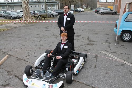 Zwei Männer in formeller Kleidung, einer sitzt in einem Go-Kart und der andere steht dahinter, auf einem Parkplatz mit geparkten Autos und einer Bank.