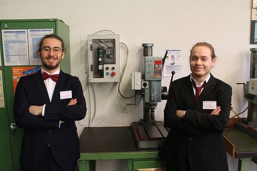 Zwei Männer stehen in einer Werkstatt mit einer Bohrmaschine und elektrischen Kästen im Hintergrund. Sie tragen Anzüge mit Fliegen und weißen Hemden.