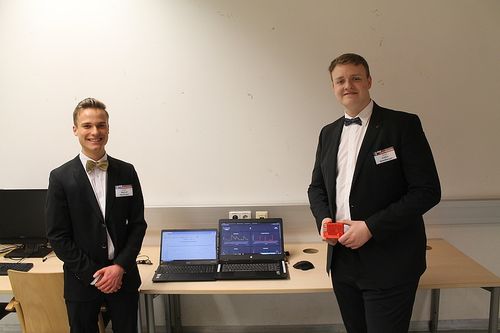 Zwei Männer in Anzügen stehen neben einem Tisch mit Laptops und einem roten Gerät. Einer hält ein rotes Gerät. Beide tragen Fliegen.