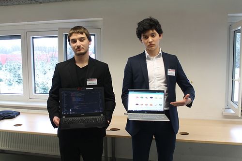 Zwei Männer in formeller Kleidung stehen in einem Raum und halten jeweils einen Laptop. Ein Laptop-Bildschirm zeigt eine Rasteransicht von Symbolen, während der andere Text auf einem schwarzen Bildschirm anzeigt. Sie stehen vor einem Schreibtisch und Fenstern.
